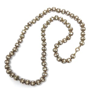 Op de afbeelding: Een vintage ketting met een reeks sierlijke, bolvormige kralen. Elke kraal is een bronzen metalen bol die sprankelende, heldere kristallen omsluit. De ketting heeft een klassiek ontwerp met een eenvoudige sluiting.