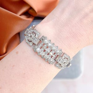 Könnte beinhalten: Ein silberfarbenes Armband mit klaren, funkelnden Strasssteinen. Das Armband hat ein geometrisches Design mit rechteckigen und kreisförmigen Steinen, die in einem detaillierten, verzierten Muster angeordnet sind. Der Verschluss ist ebenfalls mit Strasssteinen verziert.