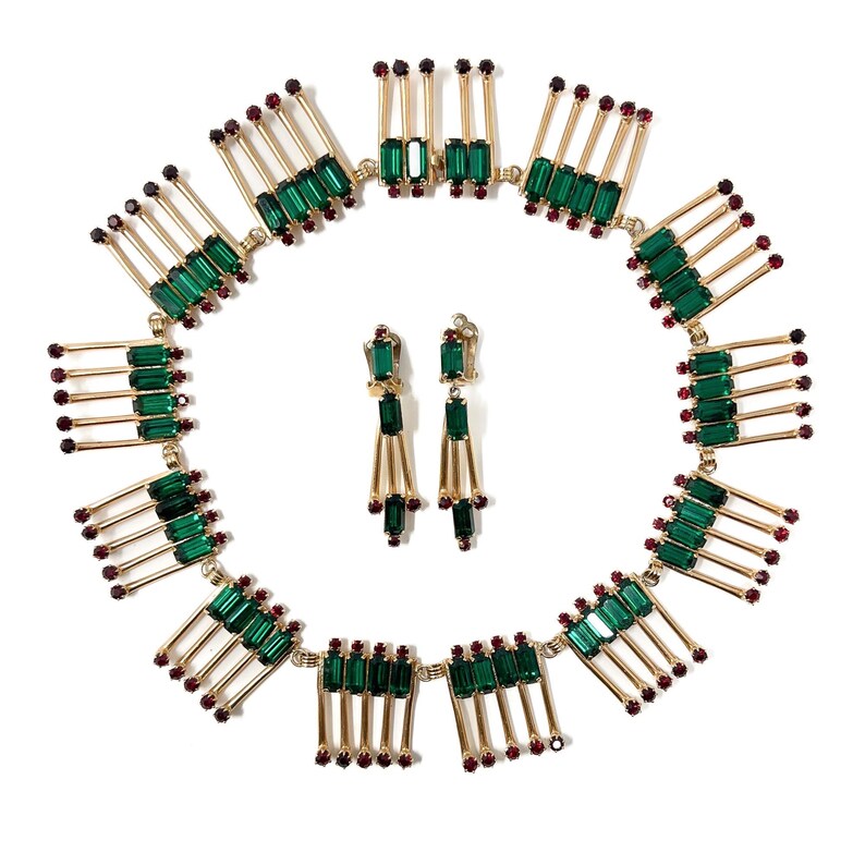 May include: A vintage jewelry set featuring a necklace and matching earrings. The necklace and earrings have a gold-tone metal base with emerald green rectangular stones and small red accents. The necklace is circular, and the earrings are dangling.