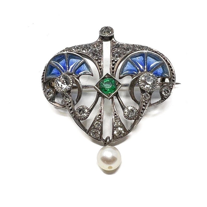 K&ouml;nnte beinhalten: Eine Brosche aus Silber und Emaille mit einem Perlenanh&auml;nger. Die Brosche zeigt ein stilisiertes Design mit blauem Emaille und einem gr&uuml;nen Edelstein.