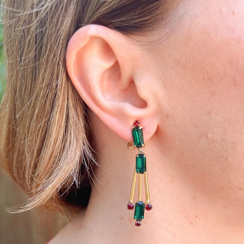 May include: Gold-toned drop earrings featuring emerald green and ruby red rectangular and round gemstones. The earrings have a linear design with a small red gem at the top and bottom, and a larger green gem in the center.