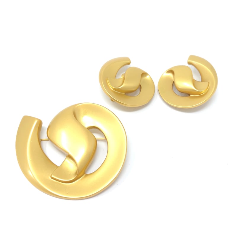 May include: A gold-toned brooch and matching earrings set featuring a stylised, abstract design. The jewellery pieces are made of a smooth, shiny metal.