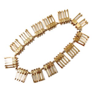 May include: A gold-toned necklace with a repeating pattern of rectangular and linear elements. The necklace is composed of multiple connected segments, each featuring a series of vertical bars. The design is symmetrical and appears to be made of metal.
