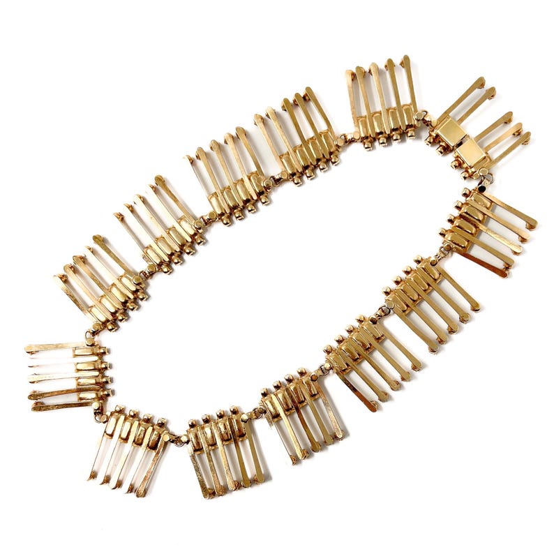 May include: A gold-toned necklace with a repeating pattern of rectangular and linear elements. The necklace is composed of multiple connected segments, each featuring a series of vertical bars. The design is symmetrical and appears to be made of metal.
