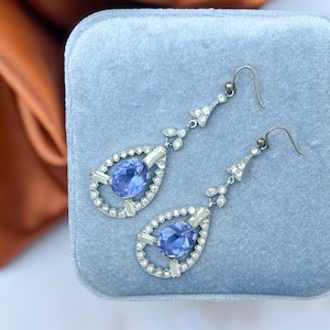 May include: A pair of silver-toned drop earrings with a blue gemstone center and clear accent stones. The earrings have a teardrop shape with a decorative top and a hook closure. The earrings are displayed on a blue velvet surface.