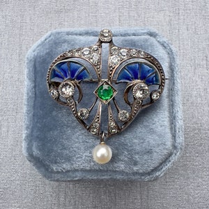 K&ouml;nnte beinhalten: Eine silberne Brosche mit herzf&ouml;rmigem Design, mit blauem Emaille, einem gr&uuml;nen Edelstein und einem Perlenanh&auml;nger. Die Brosche ist mit zahlreichen kleinen Diamanten besetzt.