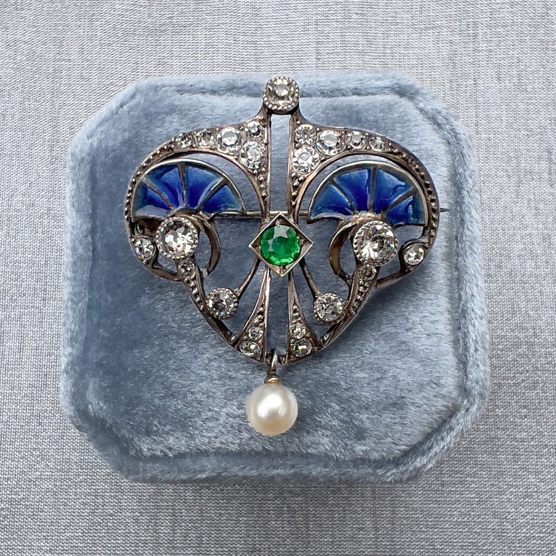 K&ouml;nnte beinhalten: Eine silberne Brosche mit herzf&ouml;rmigem Design, mit blauem Emaille, einem gr&uuml;nen Edelstein und einem Perlenanh&auml;nger. Die Brosche ist mit zahlreichen kleinen Diamanten besetzt.