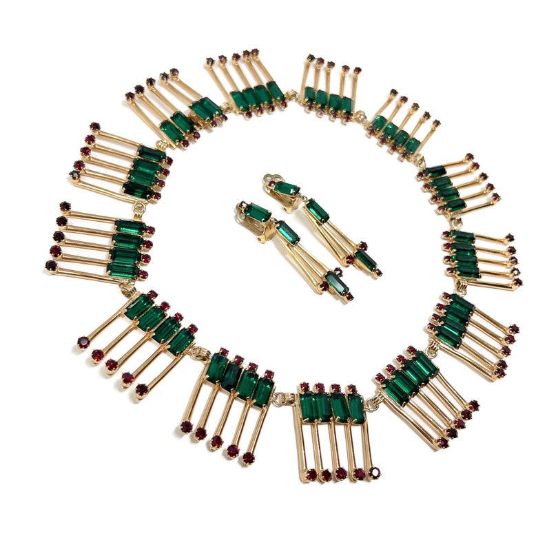 May include: A gold-toned necklace and matching earrings set. The necklace features rectangular emerald green and small ruby red gemstones. The earrings have a similar design, with emerald green and ruby red gemstones. The jewelry is set against a white background.