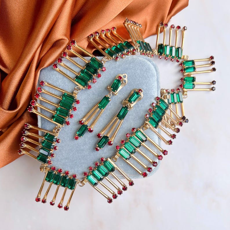 May include: A gold-toned jewelry set featuring a necklace and earrings. The necklace and earrings have a repeating design of green rectangular stones and small red stones. The jewelry is displayed on a light blue surface with a brown fabric in the background.