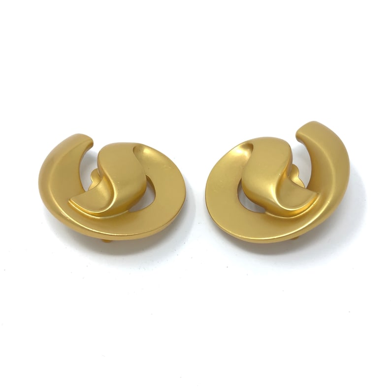 May include: A pair of gold-toned clip-on earrings with a unique abstract design. The earrings are made of a smooth, metallic material and have a slightly rounded shape.