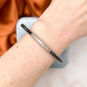 Può includere: Un braccialetto argentato con pietre rettangolari nere e trasparenti alternate. Il braccialetto è indossato su un polso, mostrando il suo design e la sua lucentezza. Le pietre sono disposte in un motivo lineare, creando un look elegante.