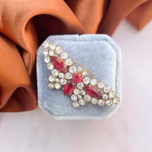 Peut inclure: Une broche en forme de papillon dorée avec des strass rouges et transparents. La broche est présentée dans un écrin en velours bleu clair.