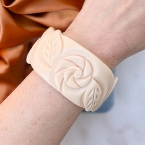 Könnte beinhalten: Cremefarbenes, breites Armband mit einem geschnitzten Blumen- und Blattdesign. Das Armband hat ein zentrales Rosenmotiv und Blattakzente. Das Armband besteht aus einem festen, undurchsichtigen Material.