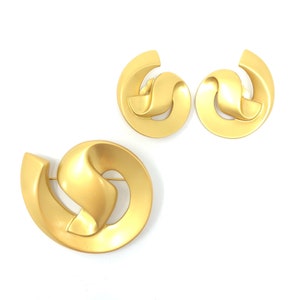 May include: A set of gold-toned earrings and a brooch with a modern abstract design. The jewellery pieces are made of a smooth, shiny metal and feature a swirling, knotted shape.
