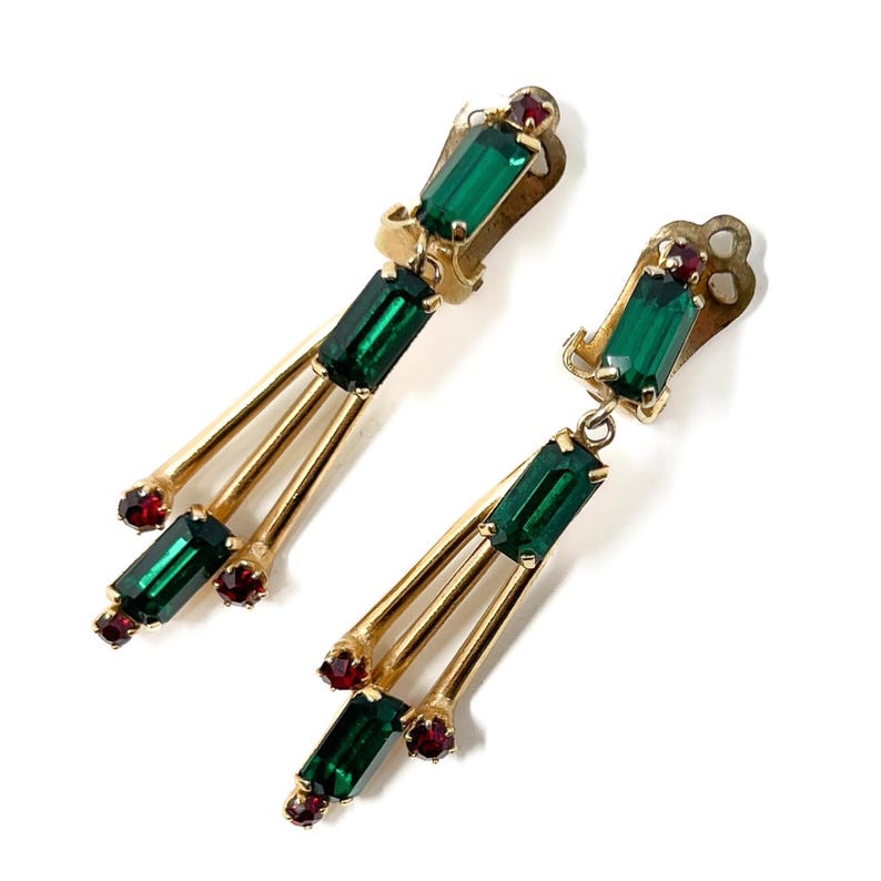 May include: A pair of vintage gold-toned earrings with a dangling design. Each earring features a clip-on top with a rectangular emerald green stone and a small red stone. Three gold bars extend downward, each ending with an emerald green and red stone.
