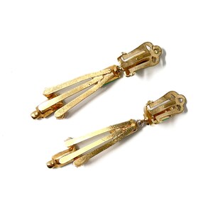 May include: A pair of vintage gold-tone clip-on earrings. Each earring features a rectangular clip, a small green accent, and three gold bars that extend downward. The earrings are set against a white background.