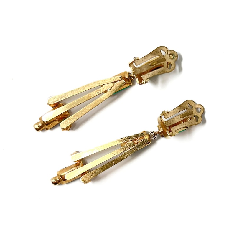 May include: A pair of vintage gold-tone clip-on earrings. Each earring features a rectangular clip, a small green accent, and three gold bars that extend downward. The earrings are set against a white background.