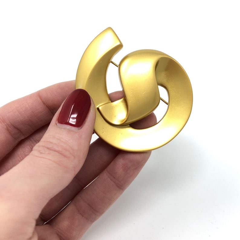 May include: A gold-toned abstract brooch with a swirling design. The brooch is made of metal and has a smooth, polished finish.