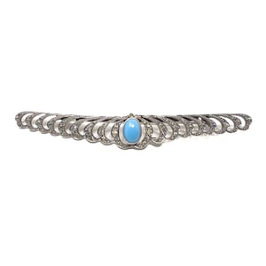 May include: A silver brooch with a blue turquoise stone in the center. The brooch is decorated with a row of small clear stones.
