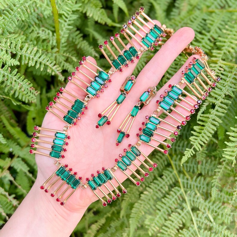 May include: A vintage jewelry set featuring a necklace and earrings. The set has gold-tone metal with emerald green and ruby red accents. The necklace and earrings have a geometric design with rectangular green stones and small red stones.