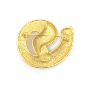 May include: A gold-toned brooch with a swirling, abstract design. The brooch has a pin and catch closure on the back.