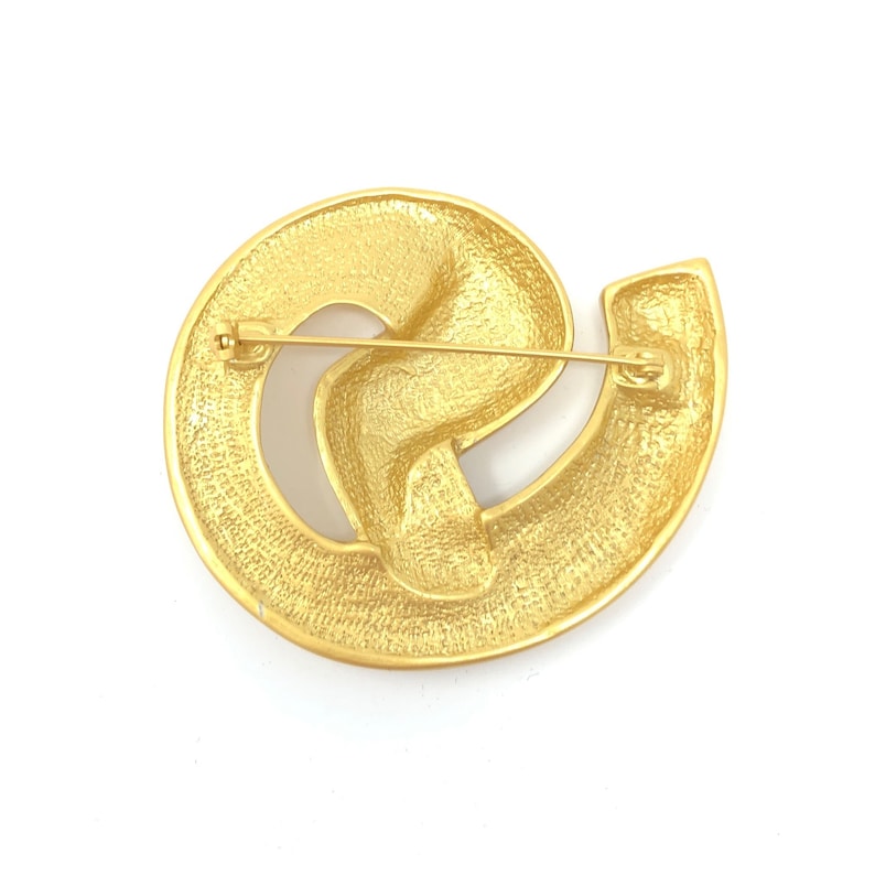 May include: A gold-toned brooch with a swirling, abstract design. The brooch has a pin and catch closure on the back.
