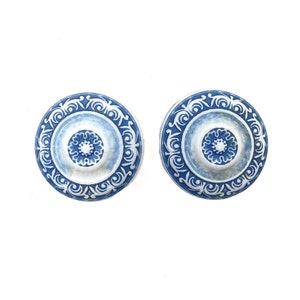 Puede incluir: Un par de pendientes de cerámica azul y blanco con un diseño circular. Los pendientes presentan un centro blanco con un patrón floral azul y un anillo exterior azul con un patrón floral blanco.