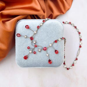 Peut inclure: Un collier argenté orné de pierres rouges et transparentes, présenté sur un écrin de velours bleu clair. Le collier présente un motif central avec des pierres rouges en forme de goutte et rondes, et une chaîne de pierres rouges et transparentes alternées.