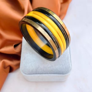 May include: A striking rectangular bangle bracelet with alternating bands of black, yellow, and gold-toned metal. The bracelet's design features several layers, creating a bold contrast. The style is modern and visually appealing.
