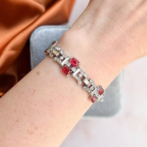 Könnte beinhalten: Ein silberfarbenes Armband mit rechteckigen roten Edelsteinen und kleinen klaren Steinen. Das Armband wird am Handgelenk getragen und zeigt sein Art-Deco-Design und seine komplizierten Details. Die Edelsteine sind in einem geometrischen Muster angeordnet.