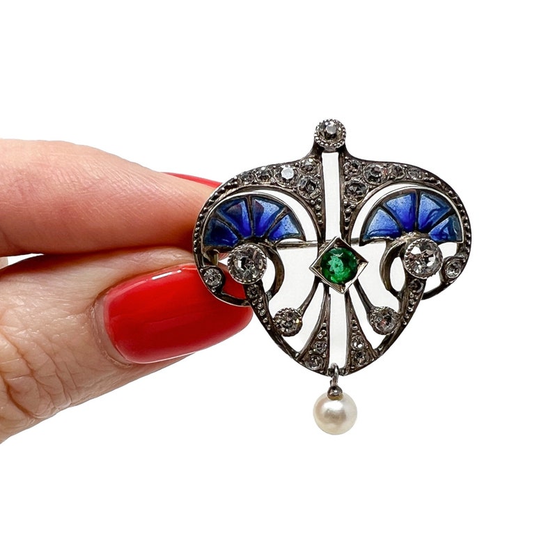 K&ouml;nnte beinhalten: Eine Brosche aus Silber und Emaille mit herzf&ouml;rmigem Design. Die Brosche ist mit blauem und gr&uuml;nem Emaille, Diamanten und einem einzelnen Perlenanh&auml;nger verziert.