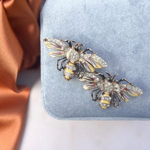 Puede incluir: Dos broches de abeja de oro con diamantes de imitación transparentes y verdes. Las abejas están sobre un fondo de terciopelo azul claro.