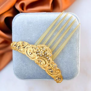 Könnte beinhalten: Ein Vintage-Haarkamm mit goldenem Filigranmuster und transparenten Zinken. Der goldene Teil ist mit kleinen, funkelnden Kristallen und floralen Mustern verziert. Der Kamm liegt auf einer hellblauen Samtoberfläche.