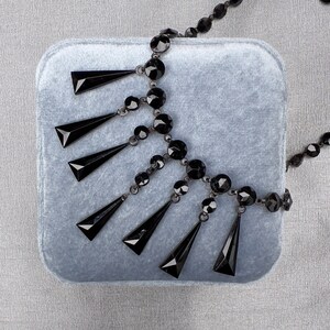 May include: A black beaded necklace with a teardrop design. The necklace is on a gray velvet background.