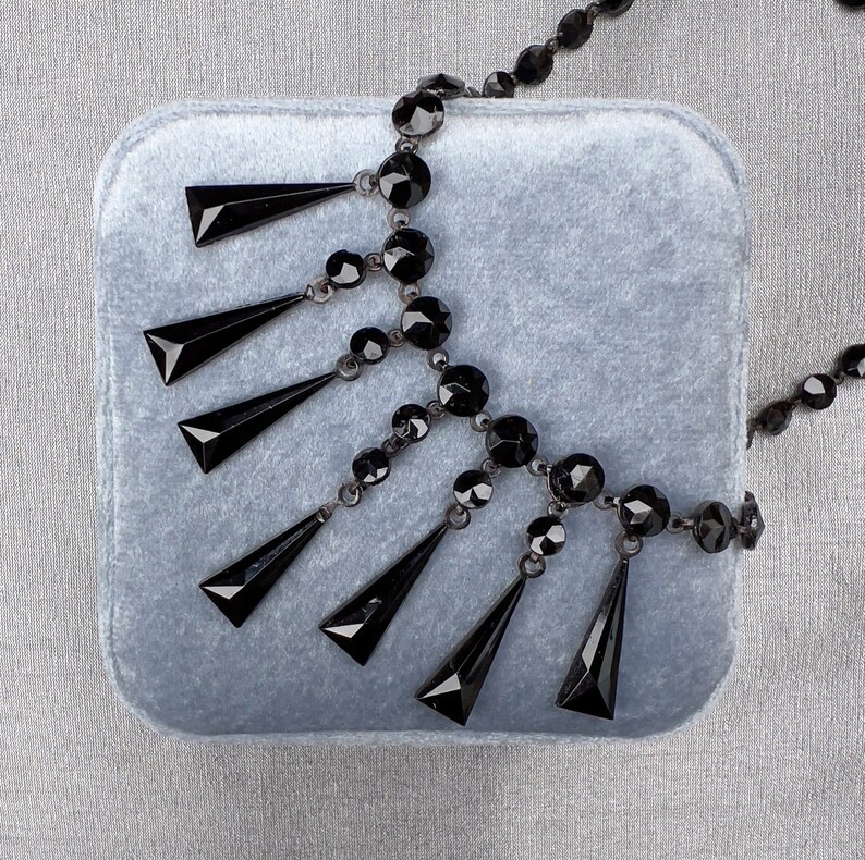 May include: A black beaded necklace with a teardrop design. The necklace is on a gray velvet background.