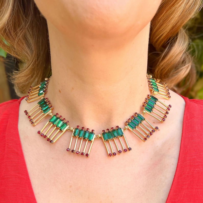 May include: A gold-toned necklace with emerald green and ruby red accents. The necklace features rectangular green stones and small red stones, arranged in a geometric pattern. The necklace is worn against a red background.