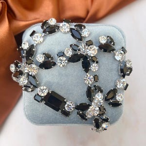 Puede incluir: Un broche de pedrería negro y blanco con un diseño floral. El broche está hecho de metal y tiene un acabado plateado.