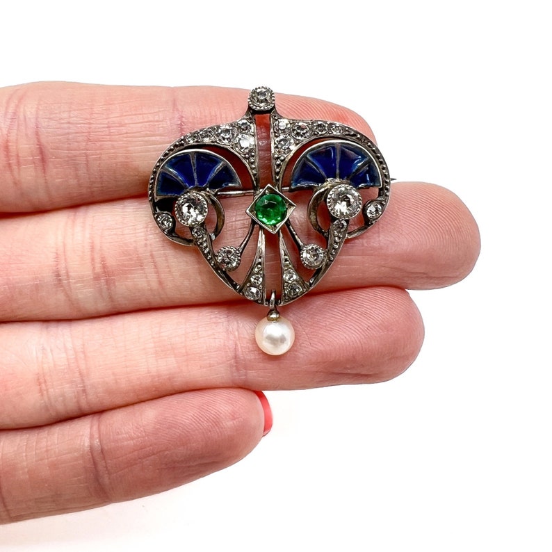 K&ouml;nnte beinhalten: Ein silberner und emaillierter Anh&auml;nger mit herzf&ouml;rmigem Design. Der Anh&auml;nger ist mit einem gr&uuml;nen Smaragd, blauem Emaille und wei&szlig;en Diamanten verziert. Eine einzelne wei&szlig;e Perle h&auml;ngt am unteren Ende des Anh&auml;ngers.