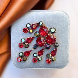 Puede incluir: Un broche vintage con un grupo de diamantes de imitación rojos e iridiscentes en un diseño en cascada. El broche está engastado en metal dorado.