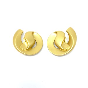 May include: A pair of gold-tone clip-on earrings with a unique, abstract design. The earrings are made of a smooth, metallic material and have a slightly rounded shape.