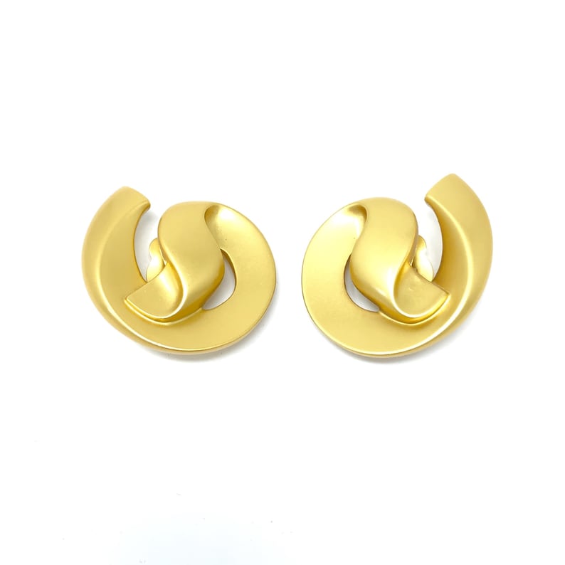 May include: A pair of gold-tone clip-on earrings with a unique, abstract design. The earrings are made of a smooth, metallic material and have a slightly rounded shape.