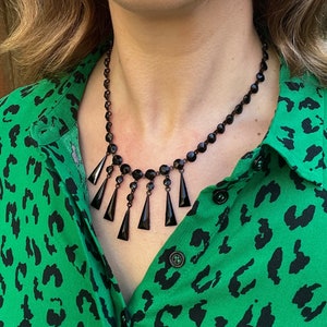 May include: A black beaded necklace with dangling teardrop-shaped crystals. The necklace is worn by a person with a green and black animal print shirt.