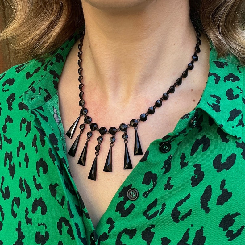 May include: A black beaded necklace with dangling teardrop-shaped crystals. The necklace is worn by a person with a green and black animal print shirt.