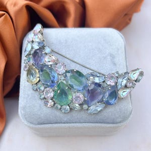 Puede incluir: Un broche en forma de media luna con un alfiler plateado, adornado con una variedad de piedras preciosas de colores. El broche presenta una mezcla de piedras azules, verdes, moradas y transparentes en varias formas y tamaños, sobre un fondo de terciopelo azul claro.