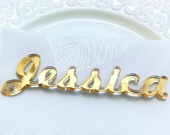 Puede incluir: Una tarjeta de lugar acrílica dorada con el nombre "Jessica" en escritura cursiva. La tarjeta de lugar descansa sobre una servilleta de lino blanca, con un plato decorativo blanco en el fondo.