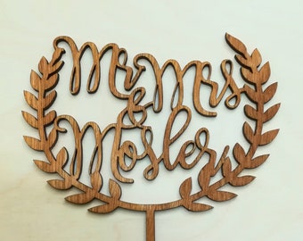 Puede incluir: Un adorno para pastel de madera con las palabras "Mr & Mrs Mosler" en fuente cursiva, rodeado por un diseño de corona de laurel. El adorno es de madera marrón claro y tiene un palillo puntiagudo para insertar en un pastel.