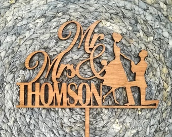 Puede incluir: Un adorno para tarta de boda de madera con las palabras "Mr & Mrs THOMSON" en una fuente decorativa. El adorno presenta siluetas de una novia y un novio. El adorno es de madera marrón claro.