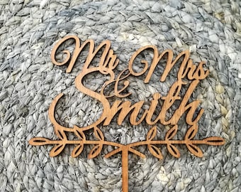 Puede incluir: Un adorno para tarta de madera con las palabras "Mr & Mrs Smith" en fuente cursiva. El adorno tiene un diseño decorativo de hojas y una estaca puntiaguda para insertar en una tarta. La madera es de color marrón claro natural.