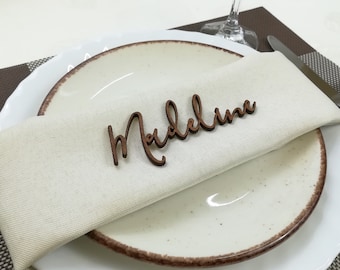 May include: A white plate with a brown rim and a beige napkin with a wooden name card that says "Madeline".
