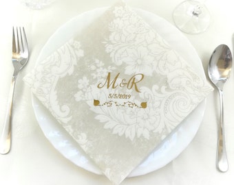 May include: A folded, square napkin with a floral pattern and gold text reading "M&R" and the date "5/5/2019". The napkin is placed on a white plate, with a fork and spoon visible.